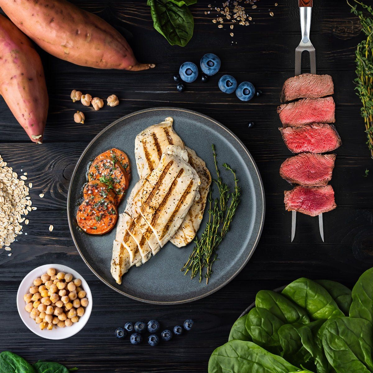 Grilled chicken, sliced beef, sweet potato, spinach, blueberries and oats laid out on a dark board